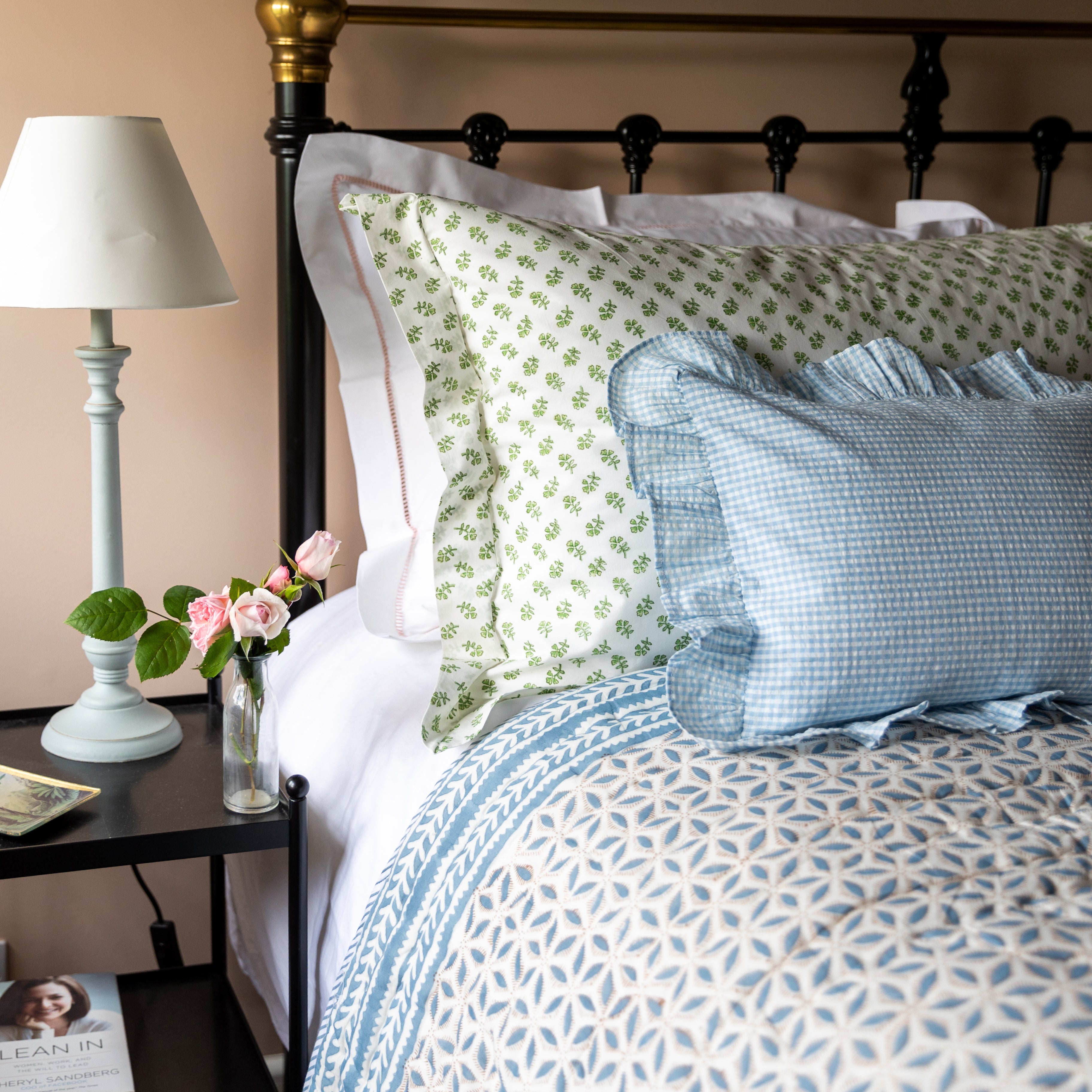 Bedroom with a bed featuring patterned bedding and pillows, a lamp on a nightstand, and a vase with flowers.