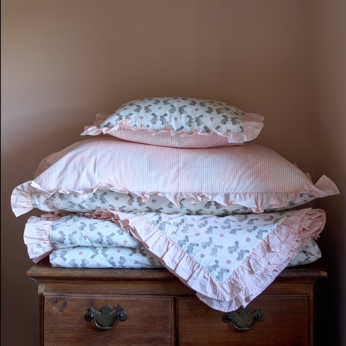 A stack of pink and floral bedding, including pillows and folded blankets with ruffled edges, displayed on a dark wooden chest against a plain brown wall.