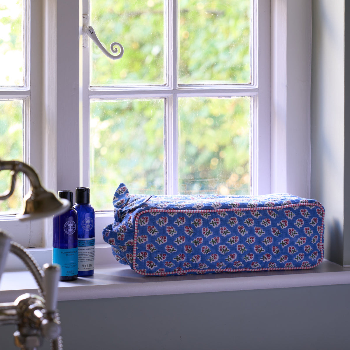 A blue floral hair accessories bag sits on a white window sill next to two blue bottles, with a window and green foliage in the background.
