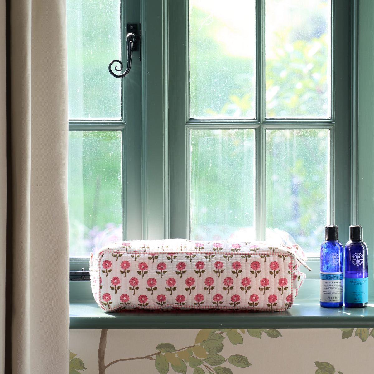 A rectangular hair accessories bag with a pink rose pattern sits on a window sill next to two blue bottles, with a green window and curtain in the background.