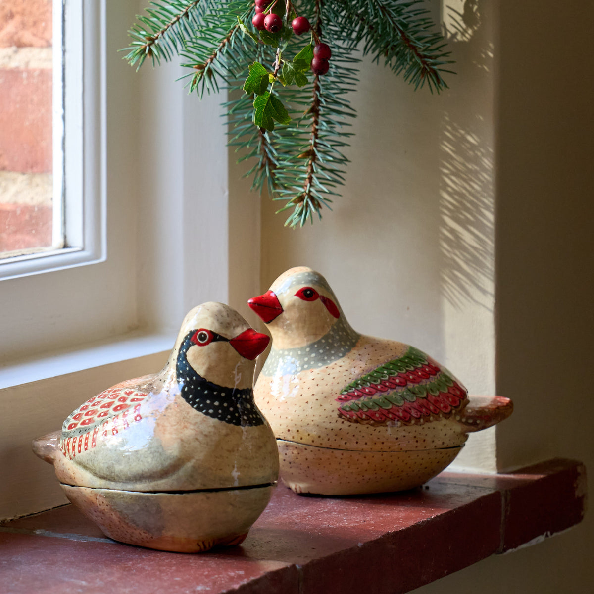 Two hand-painted papier mache patridge birds with lidded boxes sit on a terracotta window sill next to a window, with a pine branch hanging above them.